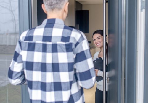 New house. Man in plaid shirt with his back to camera standing in front of open door and smiling dark-haired woman with folder inviting to house
